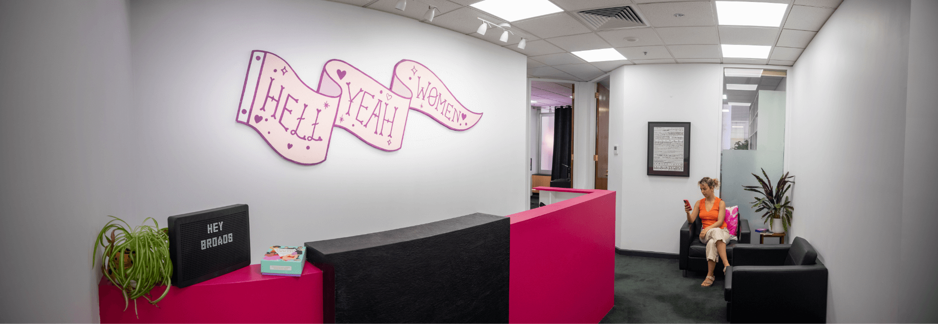A panoramic shot of the Broad Studios office with a big wall art piece that says "hell yeah girls" above the front desk